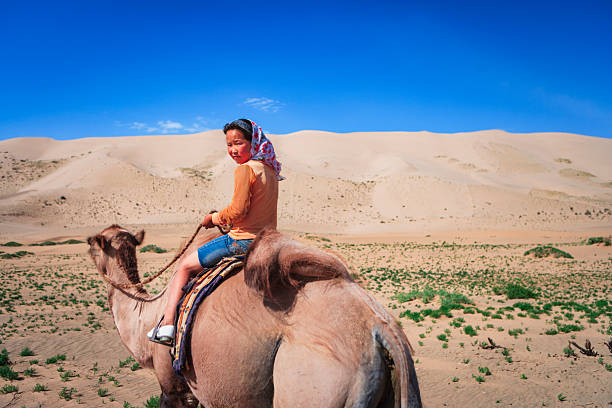 A young girl wearing a floral headscarf and orange top sits astride a camel, holding the reins and glancing back toward the camera. They traverse a sunlit sandy expanse with rolling dunes and sparse green vegetation under a vivid blue sky; a colorful saddle blanket is visible over the camel's hump. The image captures a quiet moment of traditional desert travel on a clear day.