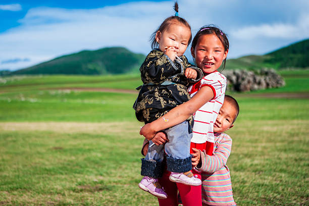Three smiling children stand on a sunlit grassy plain. An older girl holds a toddler dressed in dark, patterned traditional clothing while a younger child hugs her from the side. They wear colorful clothes and shoes, and behind them are rolling green hills under a bright blue sky with scattered clouds, creating a joyful rural scene.