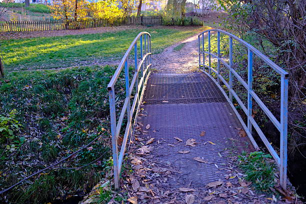 Metal pedestrian bridge with blue railings spans a small ditch, connecting a dirt path to a field.