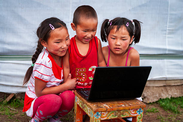 Three children sit closely together, intently watching a black laptop balanced on a small, brightly painted wooden stool. The two girls on either side wear hair clips and colorful clothing, while the younger child in the center beams with a smile. They are outdoors on grass in front of a white fabric backdrop, their faces showing curiosity and concentration as they share the screen.