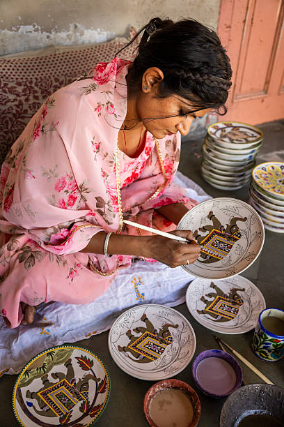 A woman in a pink floral shawl sits cross-legged on the floor, carefully painting intricate designs on round ceramic plates. Several plates with partially completed elephant motifs lie nearby, along with small bowls of colored pigments, brushes, and a stack of finished dishes. The scene focuses on the detailed handwork and traditional decorative patterns.