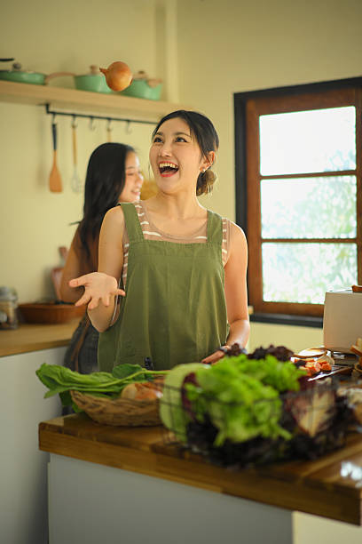 Two young women laughing while cooking together in a bright home kitchen, tossing vegetables playfully during meal preparation