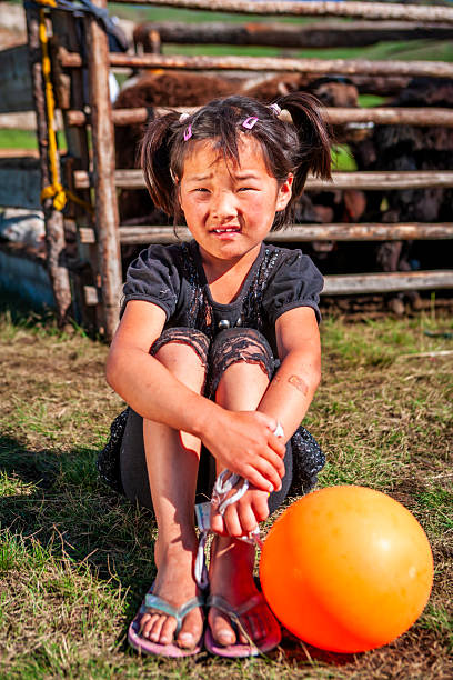 A young girl sits cross‑legged on the grass, hugging her knees and squinting in bright sunlight. She wears a black dress and lace leggings, has pigtails held by pink clips, and sandals on her feet; an orange balloon rests beside her. Behind her is a wooden corral with cattle visible, suggesting a rural outdoor setting; her face and knees show traces of dirt and a small adhesive bandage on her arm.
