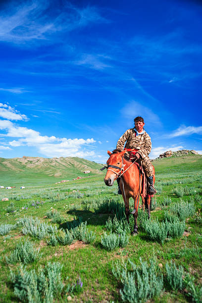 A rider sits astride a chestnut horse in the foreground of a vast grassy steppe. A brilliant, deep-blue sky with thin wisps of cloud dominates the scene, while rolling green hills and a few small white tents dot the middle distance. The rider wears an ornate patterned coat and looks toward the camera as the horse stands among low shrubs and wildflowers.