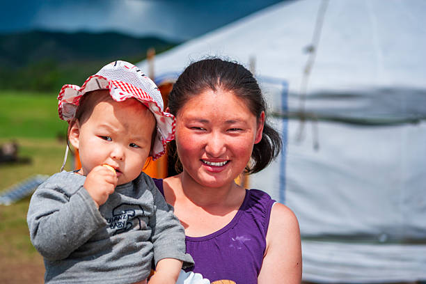 A smiling woman holds a toddler in her arms in front of a white yurt. The child wears a red-and-white sun hat and is nibbling a snack, while the woman looks at the camera with a warm expression. The scene is sunlit, set on grassy open land with blurred mountains and the yurt in the background, conveying a relaxed, rural moment.