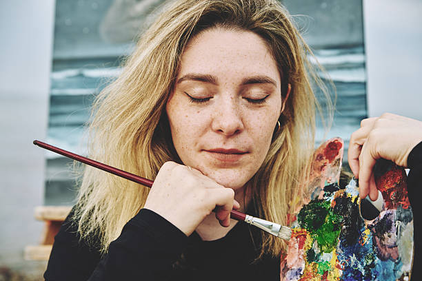 Adult woman with blonde hair and freckles holds paintbrush and colorful palette while focusing deeply on her artwork in bright studio lighting. Calm, creative moment captured with neutral background