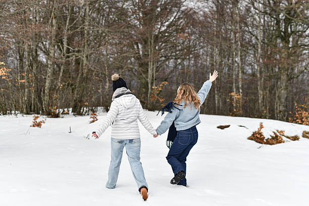 Wrapped in warm winter clothing, two friends hold hands while laughing and enjoying time outdoors in fresh snow. A warm friendship moment.