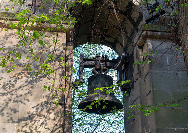 Large dark bronze church bell suspended under a weathered stone archway, framed by budding spring branches. атмосферное heritage detail for history, religion, architecture, and tradition concepts.