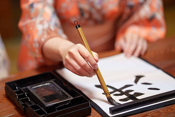 Top view of two women wearing traditional kimono practicing Japanese calligraphy with brush and black ink on white paper. Ink set placed on a wooden table in a traditional Japanese interior.