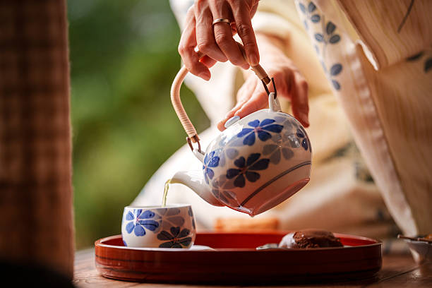 Close up of a woman wearing a traditional Japanese kimono pouring tea from a ceramic teapot into a small cup on a wooden table.