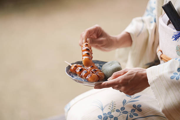 Woman wearing a traditional Japanese kimono holding a small plate with dango skewers and mochi sweets while sitting outdoors.