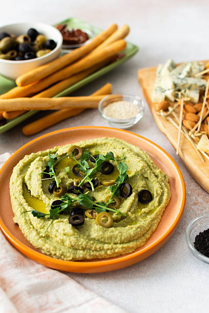 Broccoli hummus is artistically displayed in a bowl, garnished with olives. Accompanied by a charcuterie board featuring cheese, nuts, and breadsticks for a delightful snack experience