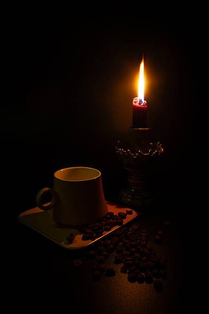 High angle view of a ceramic cup and scattered roasted coffee beans on a table, illuminated only by a single burning candle. The scene evokes a sense of peace, solitude, and warm atmosphere during night time or early morning.