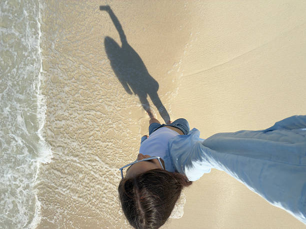 Young woman walking along wet sandy shoreline at ocean's edge, casting a long shadow under bright sunlight on tranquil beach. Woman taking selfie on vacation.