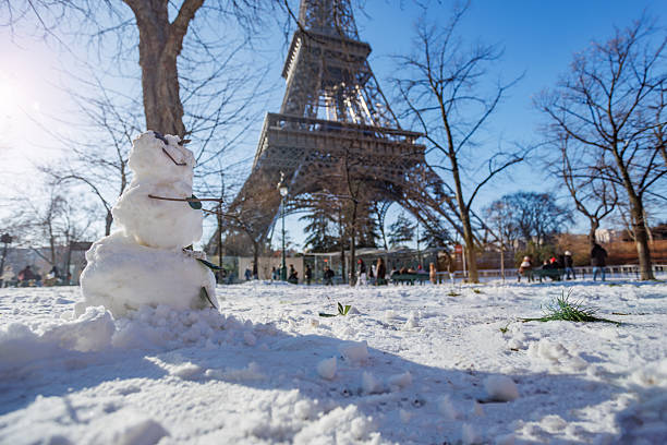 A cheerful snowman stands in the foreground with the Eiffel Tower towering in the background under a clear blue sky, surrounded by snow and bare trees in a winter park setting, France, Paris