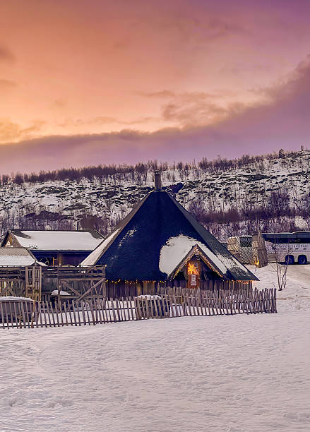Kirkenes, Norway - December 25, 2025: Snowhotel and resort. Pitch black Sami-style architecture of restaurant building with central chimney. Snow and tree-covered hill in back under noon orange cloudscape