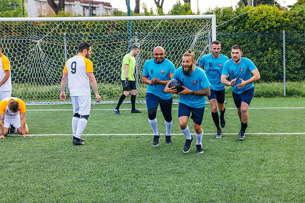 A women's football team featuring hijabi and non-hijabi players is captured during a free kick situation on the field. Players in red jerseys form a defensive wall in front of the goal, bracing for impact, while an opponent in an orange jersey prepares to take the shot.