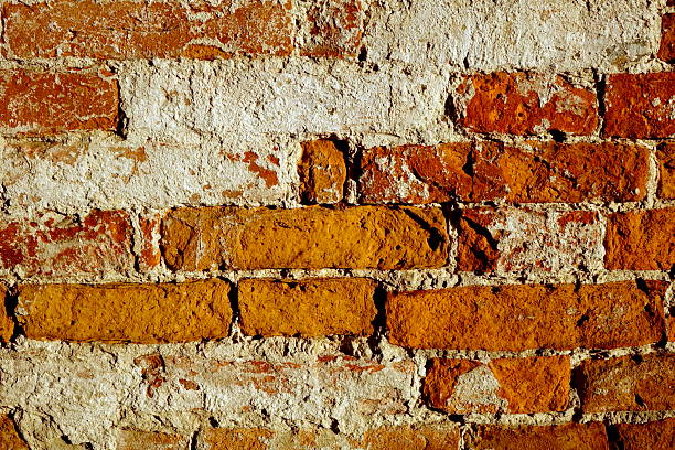 Red brick wall displays rough texture with colors of orange and red. It stands in an urban area under bright sunlight, highlighting its aged look.