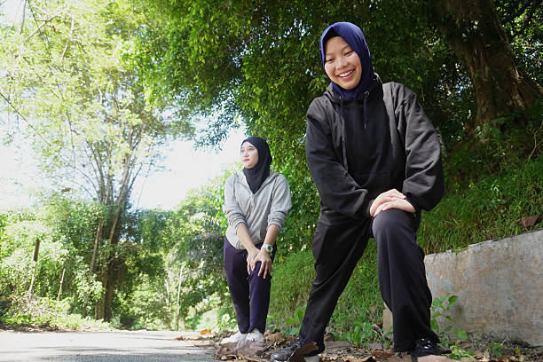 Two young Muslim women wearing hijab stretching and warming up during outdoor exercise on a sunny day in a green park. Healthy lifestyle, friendship, and fitness concept with natural light