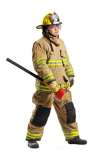 Full body firefighter wearing protective turnout gear, helmet, gloves, and boots stands holding a rescue axe. Emergency responder is isolated on a white background with firefighting equipment visible.