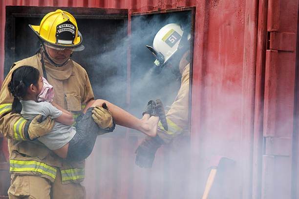 Firefighter in protective turnout gear carries child out of smoke filled container while another firefighter assists from inside. Scene shows emergency rescue operation during fire training or evacuation scenario.