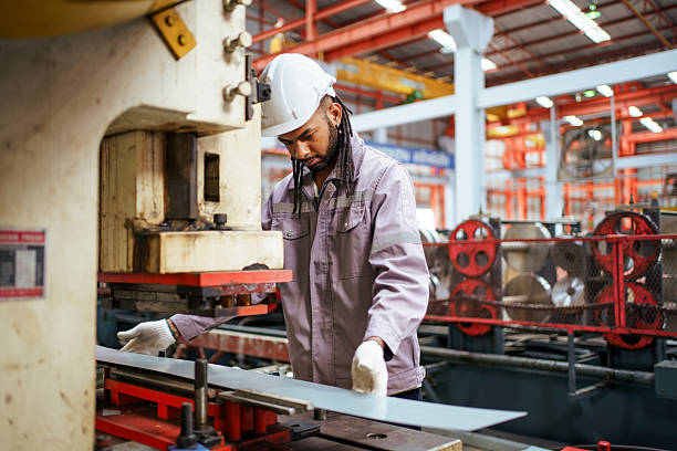 Factory worker wearing safety helmet and gloves operates metal press machine while positioning sheet metal during forming process inside manufacturing facility with industrial equipment and production line.
