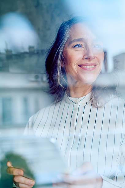 Mature woman smiling looking out window holding tablet
