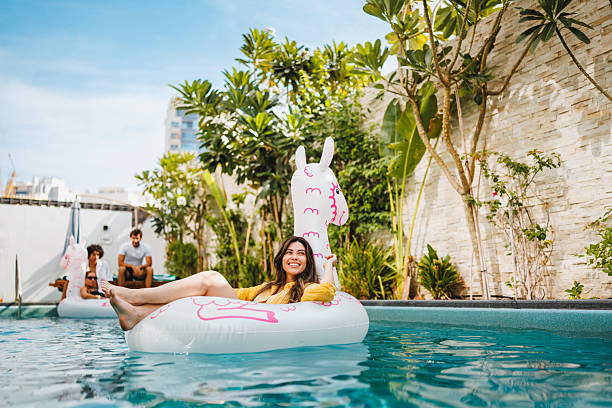 Young Hispanic female relaxing on a llama inflatable pool float in the private backyard pool. Enjoying the summer sun, she is smiling and enjoying the water. Lush green plants surround the pool under a clear blue sky.