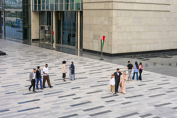 Diverse group of people are walking on a patterned plaza pavement in front of a modern glass building featuring the UAE flag, showing urban life and business activity