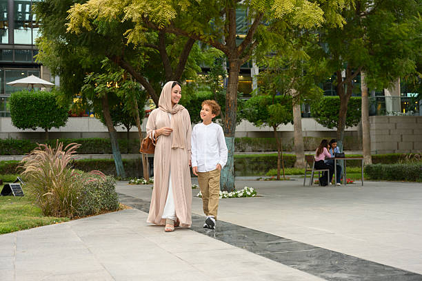 Middle Eastern mother and son in hijab and abaya stroll through a modern urban park, smiling and enjoying warm daytime bonding amid trees and green space, carefree togetherness