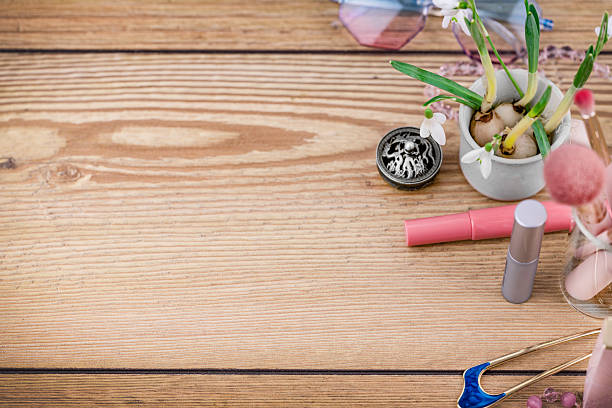 Angled tabletop still life of beauty and fashion accessories arranged on a rustic wooden table with wide copy space. Spring snowdrop flowers in a small pot, makeup items, cosmetic brushes, sunglasses, and feminine lifestyle details create a soft seasonal background for beauty, skincare, wellness, and fashion design concepts.