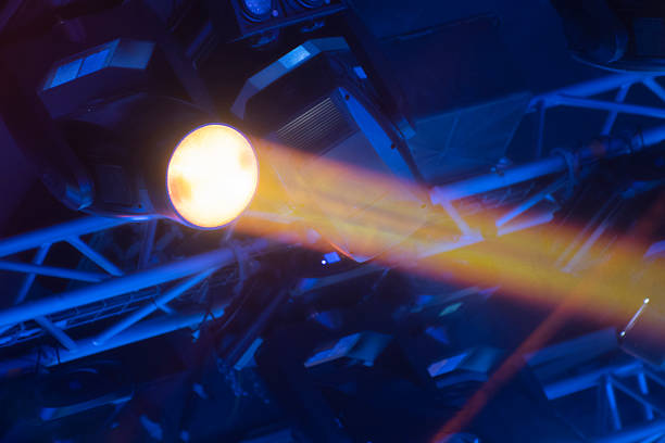 Close-up of a professional moving head spotlight on a metal truss system during a live event. Bright orange light beam against blue background. Concept of technical event production and stage lighting equipment