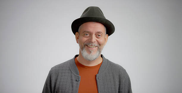Mature Caucasian male subject smiling directly at the camera in a professional studio environment. He is wearing a dark hat and a light gray cardigan over an orange shirt. The background is a clean, neutral light gray color, creating a bright and high-key lighting setup. The portrait conveys genuine happiness and approachability.