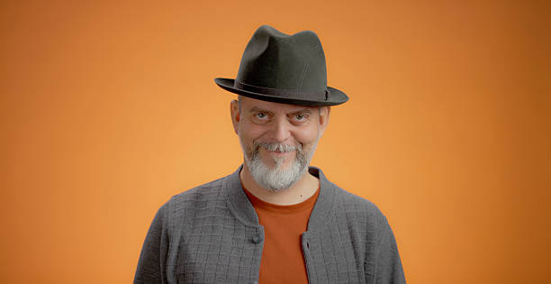A mature Caucasian man with a beard is standing in a bright studio setting, wearing a gray hat and orange jacket. He is looking directly at the camera with a neutral expression, conveying a sense of professionalism and confidence.