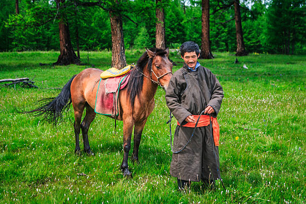 A man stands in a lush green meadow holding the reins of a saddled brown horse. He wears a long dark coat tied with a bright orange sash and smiles gently while the horse grazes nearby. The scene is framed by tall conifer trees and scattered wildflowers, creating a peaceful, pastoral atmosphere.