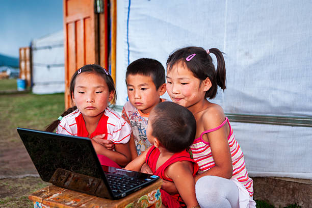 Four children sit at the open doorway of a yurt, huddled closely around a laptop placed on a small painted wooden stool. Two girls with hair clips, a boy and a toddler watch the screen intently; the older girl cradles the youngest. Soft natural light illuminates their faces and the white felt walls of the yurt, with grassy steppe and another yurt visible just outside.