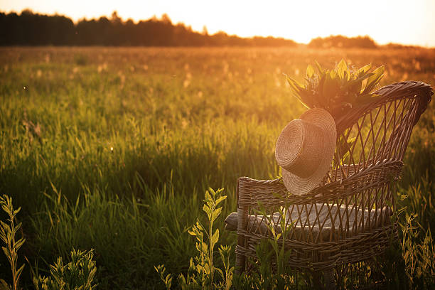 A rustic wicker chair adorned with a straw hat and wildflowers sits in a golden field as the sun sets, casting a warm glow over the scene