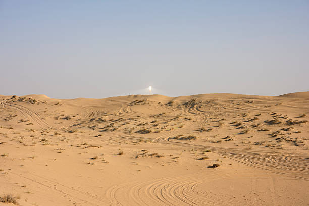 Radiant solar power tower generating clean energy on a clear sunny day, standing tall amidst arid desert landscapes with vehicle tracks on sand dunes. United Arab Emirates