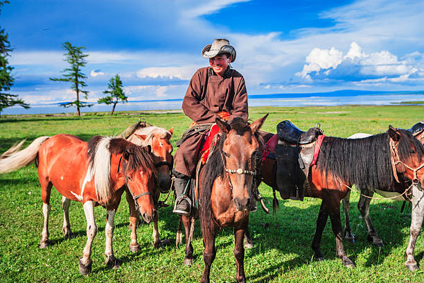 A smiling rider in traditional clothing and a cowboy-style hat sits astride a saddled horse, surrounded by several other horses grazing on a lush green meadow. Behind them a calm lake and a few scattered trees stretch to the horizon beneath a wide blue sky dotted with clouds, creating a bright, peaceful pastoral scene.