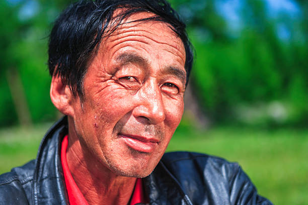 Close-up color portrait of a middle-aged man outdoors. He wears a black leather jacket and red shirt, with weathered skin, fine wrinkles around his eyes, a slight smile, and a calm, thoughtful expression against a soft green blurred background.
