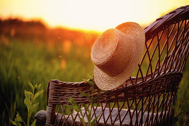 A woven straw hat is placed on a rustic wicker chair, bathed in the warm glow of a setting sun over a grassy field