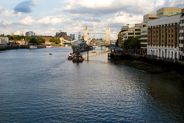 London's river thames flowing past historic landmarks including tower bridge, the tower of london, and hms belfast, creating a vibrant urban waterscape under a cloudy sky