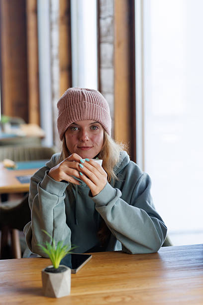 Young woman in a cozy cafe enjoying a warm drink while sitting at a wooden table with a small plant and a mobile phone nearby