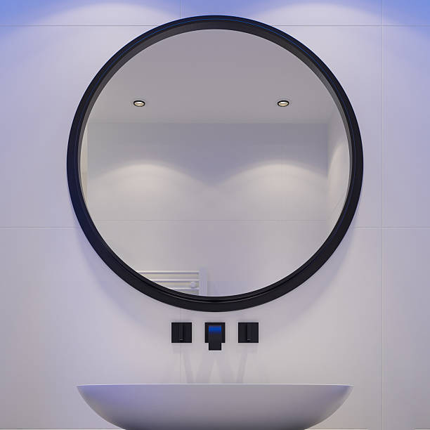 Close-up view of a contemporary minimalist bathroom featuring a round black-framed mirror mounted on large-format gray stone tiles. Below it, a sleek floating vanity cabinet with clean handleless fronts supports a white vessel sink placed on a modern countertop. Subtle ambient lighting enhances the textures and creates a calm, refined atmosphere. Ideal for use in interior design inspiration, bathroom renovation concepts, modern home decor, architectural presentations, real estate marketing, and luxury residential design content.