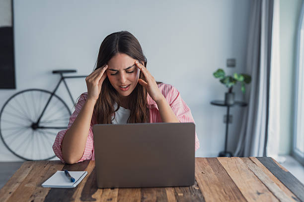Tired woman massaging temples, suffering from headache after computer work, sitting at desk with laptop at home, exhausted young female with closed eyes touching head, relieving pain, migraine