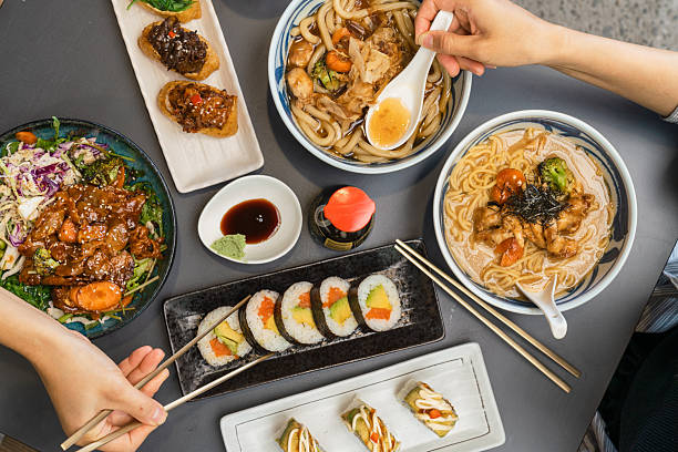 Hands picking food from a colorful Asian spread, Sushi rolls and noodles.