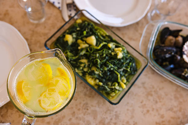 Glass pitcher of homemade lemonade with lemon slices sits alongside Mediterranean dishes on a tan stone table as a diverse multi generational family gathers for a casual outdoor vacation meal.