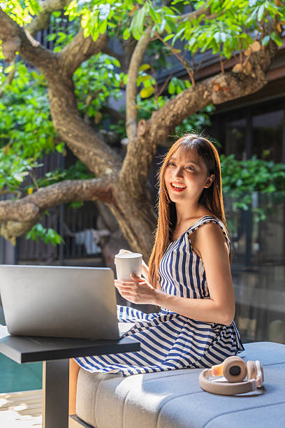 Asian woman working remotely on laptop beside luxury courtyard pool surrounded by tropical tree sunlight reflection modern architecture peaceful lifestyle digital nomad relaxation work life balance serene outdoor workspace.