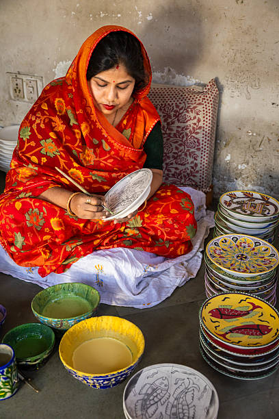 A woman wearing a bright red floral sari sits cross-legged on the floor, intently painting a detailed design onto a ceramic plate with a fine brush. Bowls of yellow and green paint are placed in front of her, and neat stacks of colorful, hand-painted plates featuring floral and fish motifs are arranged to her side. The scene shows a modest indoor workspace with a patterned cushion and a worn wall in the background, highlighting traditional handcraft work.