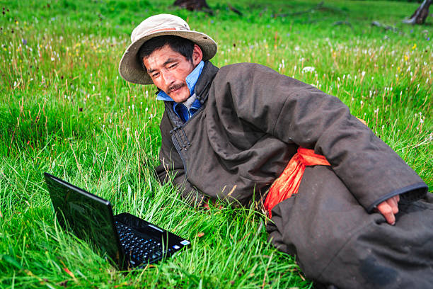 A man wearing a wide-brimmed hat and a heavy, warm coat with a bright orange sash lies on lush green grass, intently looking at a small open laptop set in front of him. The scene shows tall meadow grasses and scattered wildflowers, creating a vivid contrast between traditional rural clothing and modern technology. The mood is quiet and contemplative, evoking everyday life in a pastoral landscape.